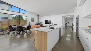 A modern kitchen with a central island, a dining area with chairs, and a view of the outdoors. - Amongst The Tea Trees (Inverloch)