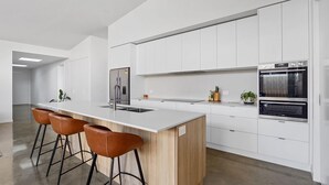 A modern kitchen with white cabinetry, a stainless steel refrigerator, an oven, and a wooden island with bar stools. - Amongst The Tea Trees (Inverloch)