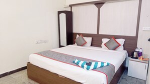 Uma cama bem arrumada com cabeceira de madeira, roupa de cama branca e um cobertor xadrez. Há uma pequena mesa de cabeceira com uma chaleira e garrafas de água.