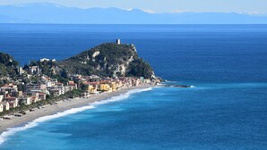 A coastal town with buildings along the beach, a prominent hill with a tower, and a clear view of the sea. - La Gioiosa (Finale Ligure)