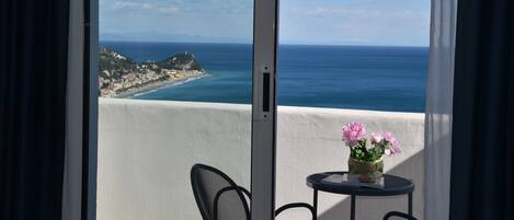 Un balcón con vistas al mar y una pequeña isla. Hay una mesa con sillas y un florero con flores.