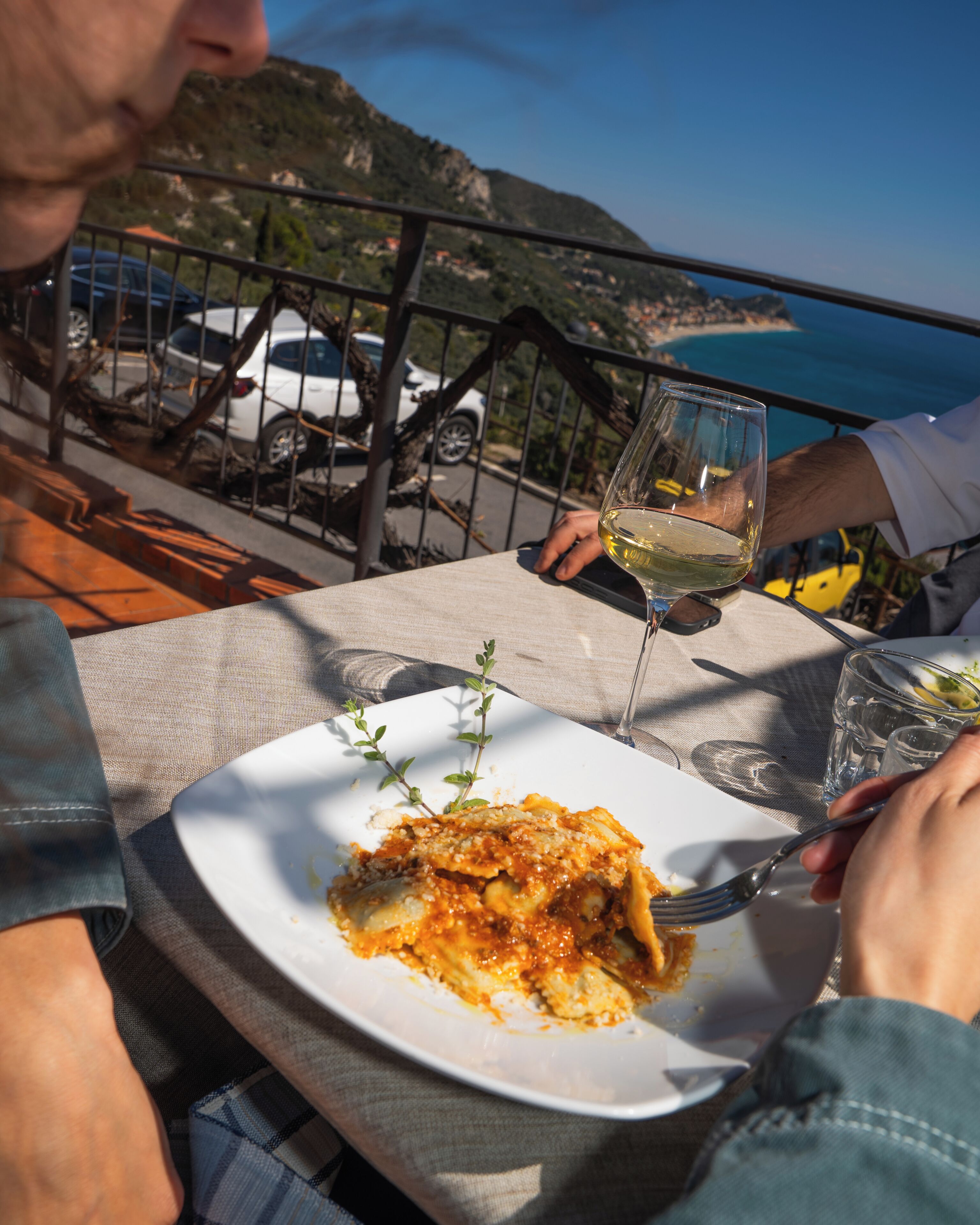 A dining table with a plate of food, a glass of wine, and a scenic view of the sea and mountains in the background.