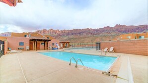 A pool area with a view of red rock formations, a covered seating area, and a building with a red roof. - Moab Mirage Hot Tub and Pool (Moab)