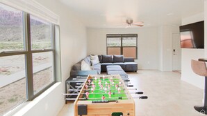A spacious living room with a foosball table, a large window offering a view of a mountainous landscape, a grey sectional sofa, and a ceiling fan. - Moab Mirage Hot Tub and Pool (Moab)