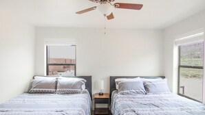 A bedroom with two beds, a ceiling fan, a nightstand with a lamp, and a window with a view of buildings. - Moab Mirage Hot Tub and Pool (Moab)