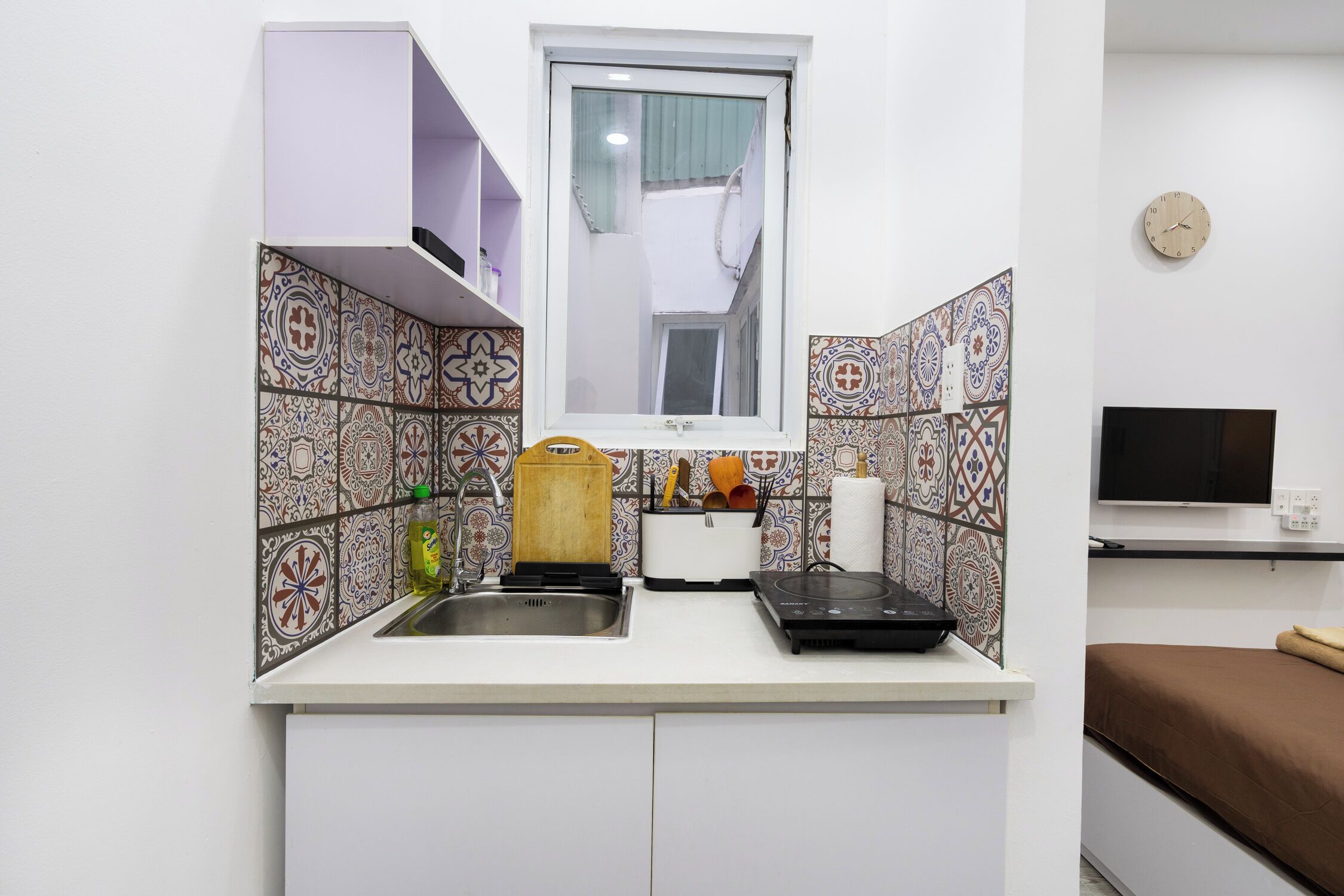 A compact kitchen with a sink, a toaster, and a microwave. There is a window above the sink and a TV mounted on the wall to the right.