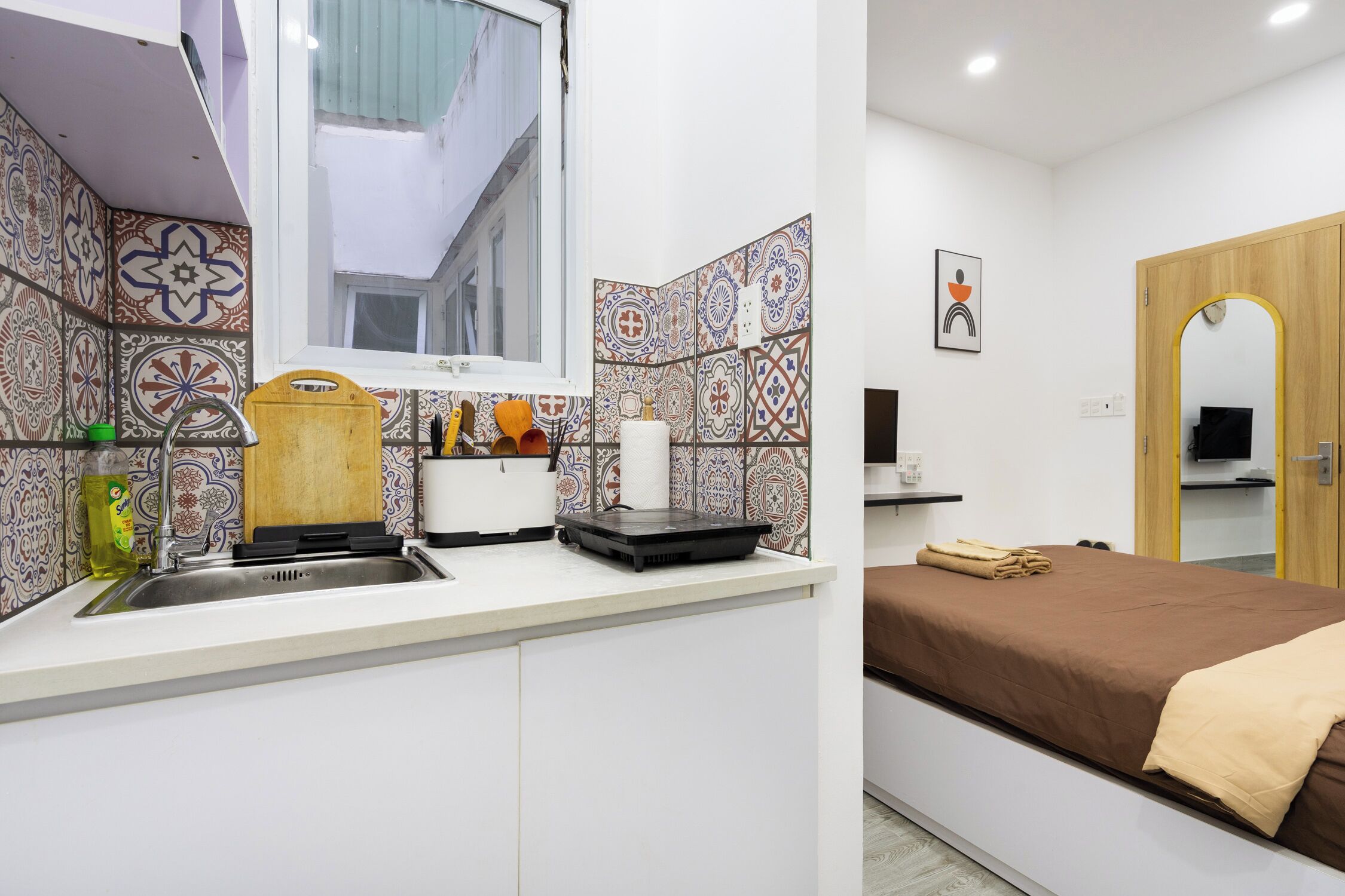 Habitación de hotel pequeña con kitchenette, una cama con cubrecama marrón y una puerta de madera.