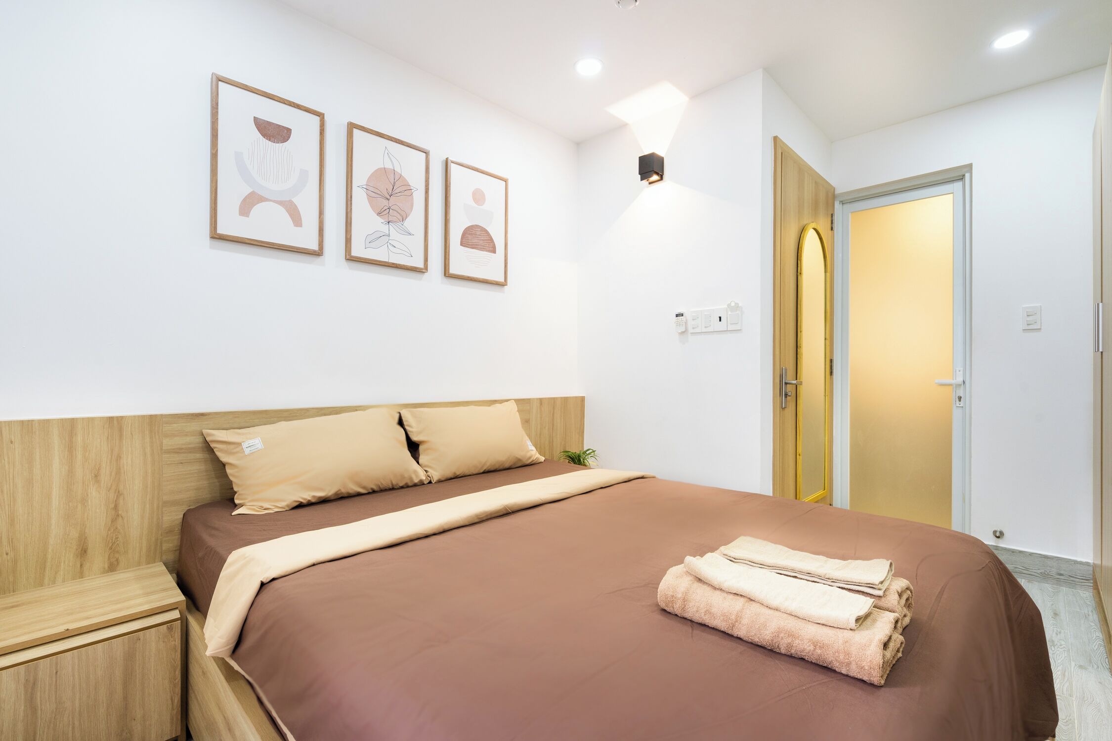 A neatly made bed with a brown bedspread and pillows, a bedside table, and three framed artworks on the wall.