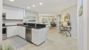 A modern kitchen with white cabinets, a black countertop, and stainless steel appliances. There is a dining area with a table and chairs, and a living room with a sofa and a lamp.