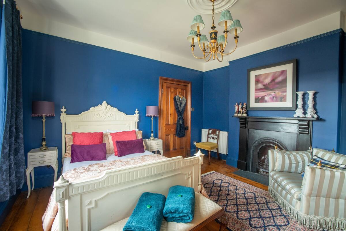 A bedroom with a white ornate bed, a blue wall, a fireplace, a striped armchair, and a chandelier.
