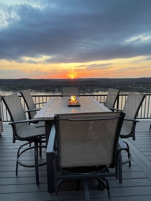 A deck with a table and chairs, overlooking a body of water and a sunset. - Amazing Sunset Lake Views, Huge Deck & Hot Tub! (Sunrise Beach)