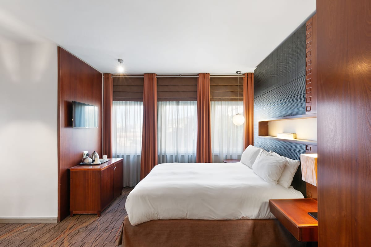 Habitación de hotel con una cama grande, una mesita de noche de madera, un televisor y una ventana con cortinas.