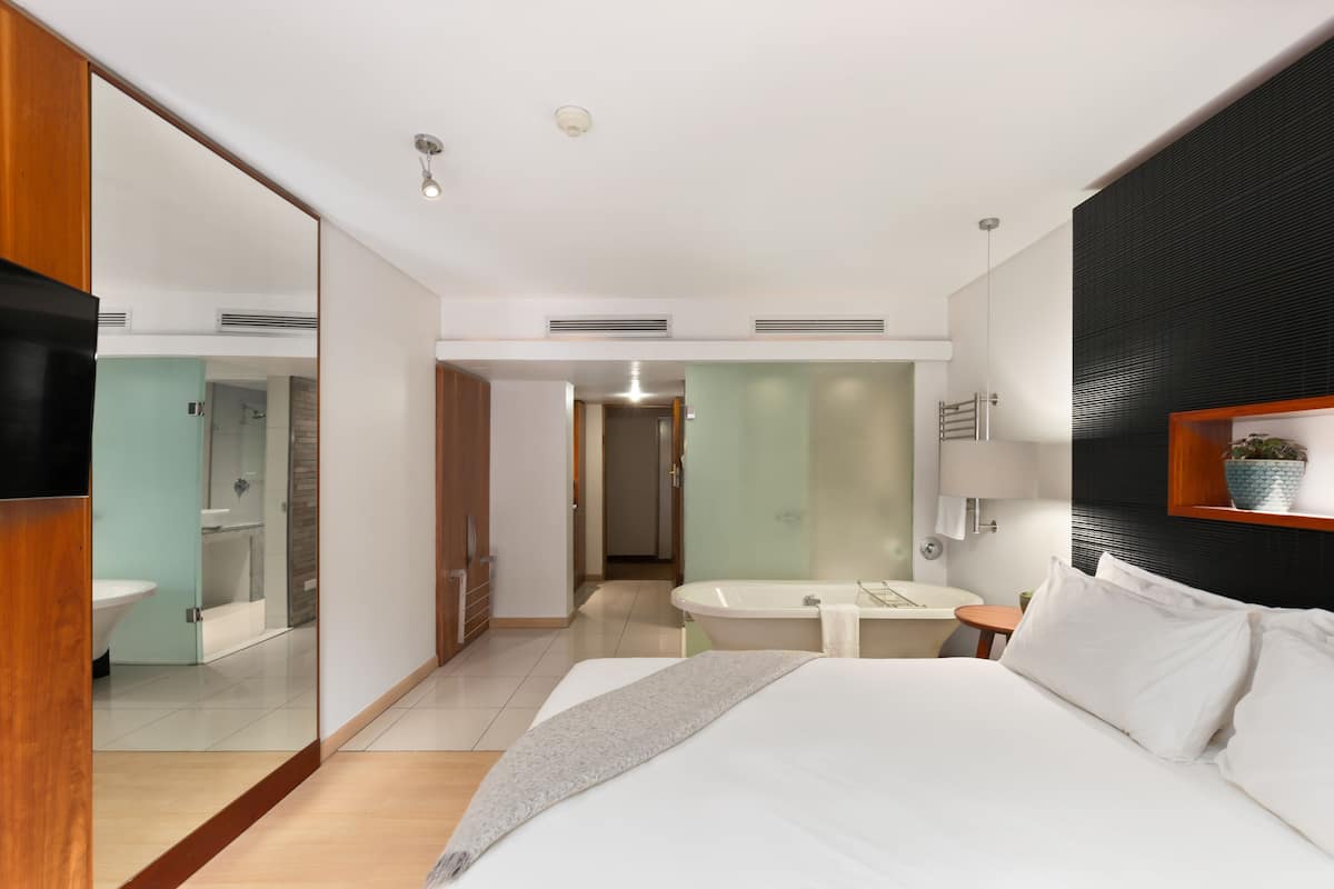 Una habitación de hotel moderna con una cama grande, una mesita de noche, un espejo y un baño con bañera y ducha.