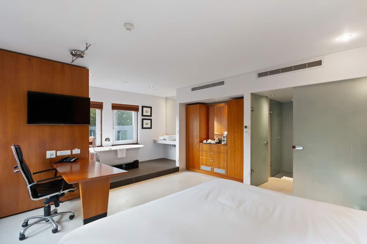 Una habitación de hotel moderna con una cama grande, un escritorio con silla, un televisor de pantalla plana, un armario y un baño con lavamanos y espejo.