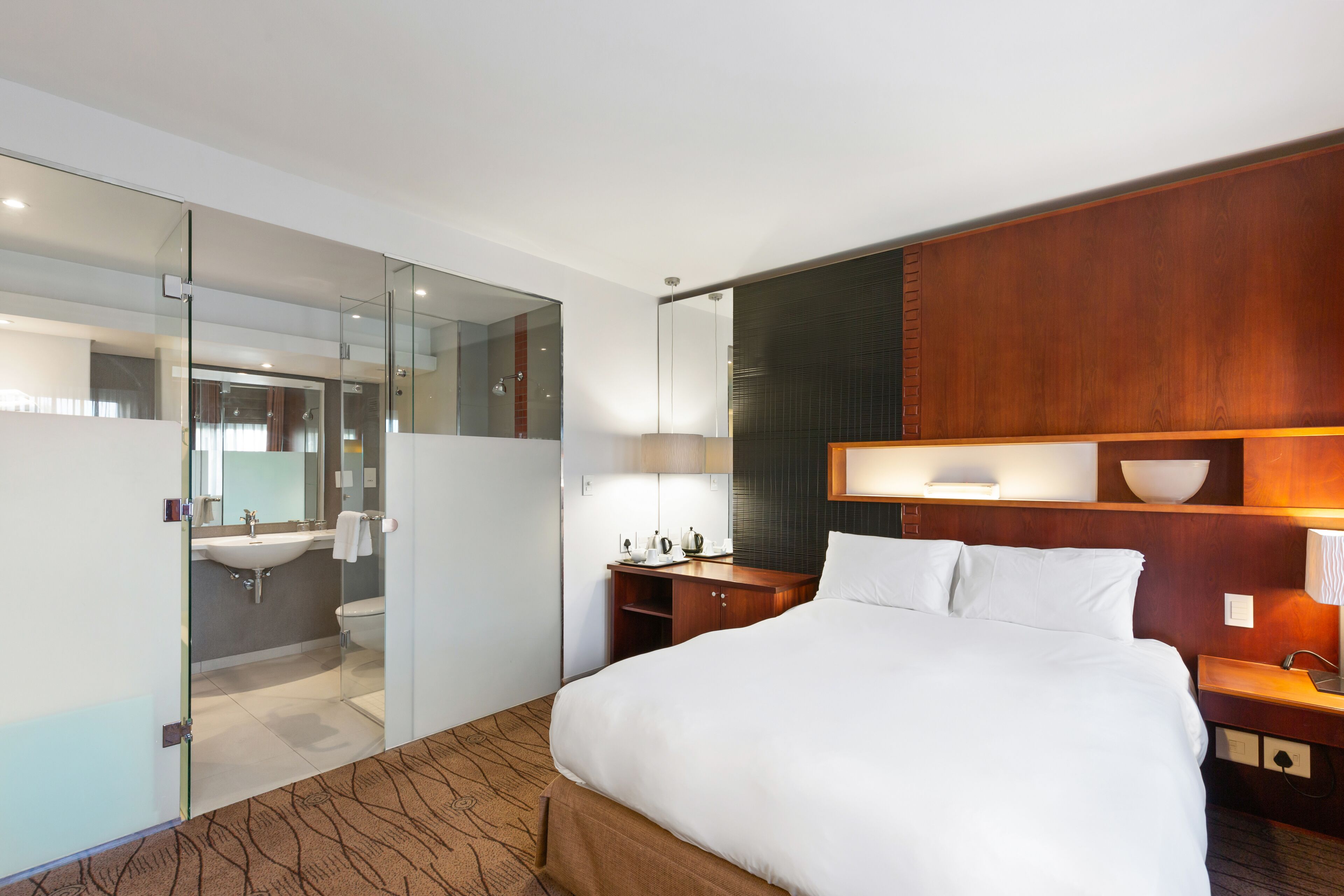 Une chambre d’hôtel moderne dotée d’un grand lit, d’une tête de lit en bois, d’une table de chevet avec une lampe, d’une commode et d’une salle de bain fermée par une paroi en verre, comprenant un lavabo et un miroir.