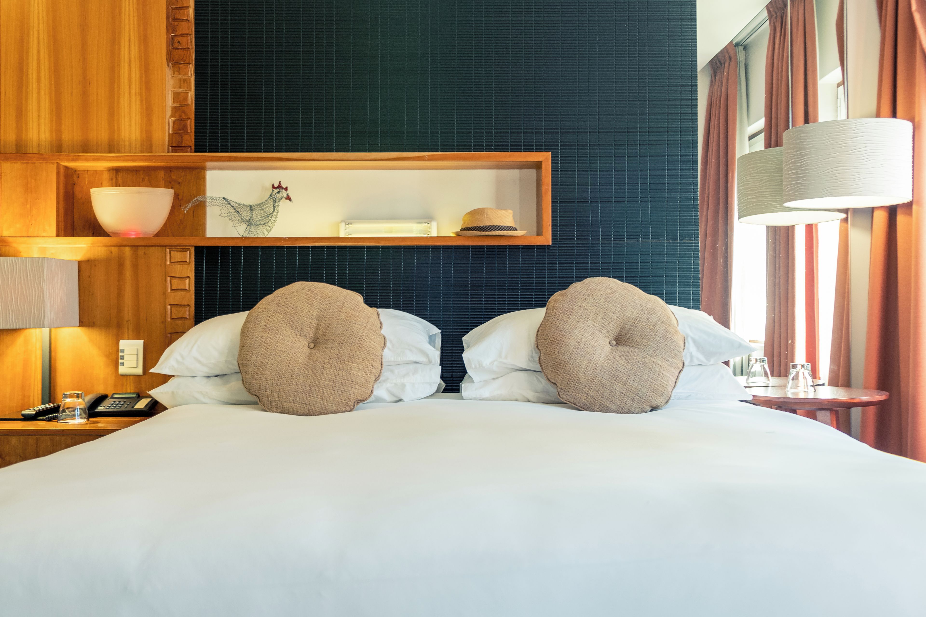 Habitación de hotel con cama, dos almohadas, mesita de noche, lámpara, espejo y repisa con objetos decorativos.