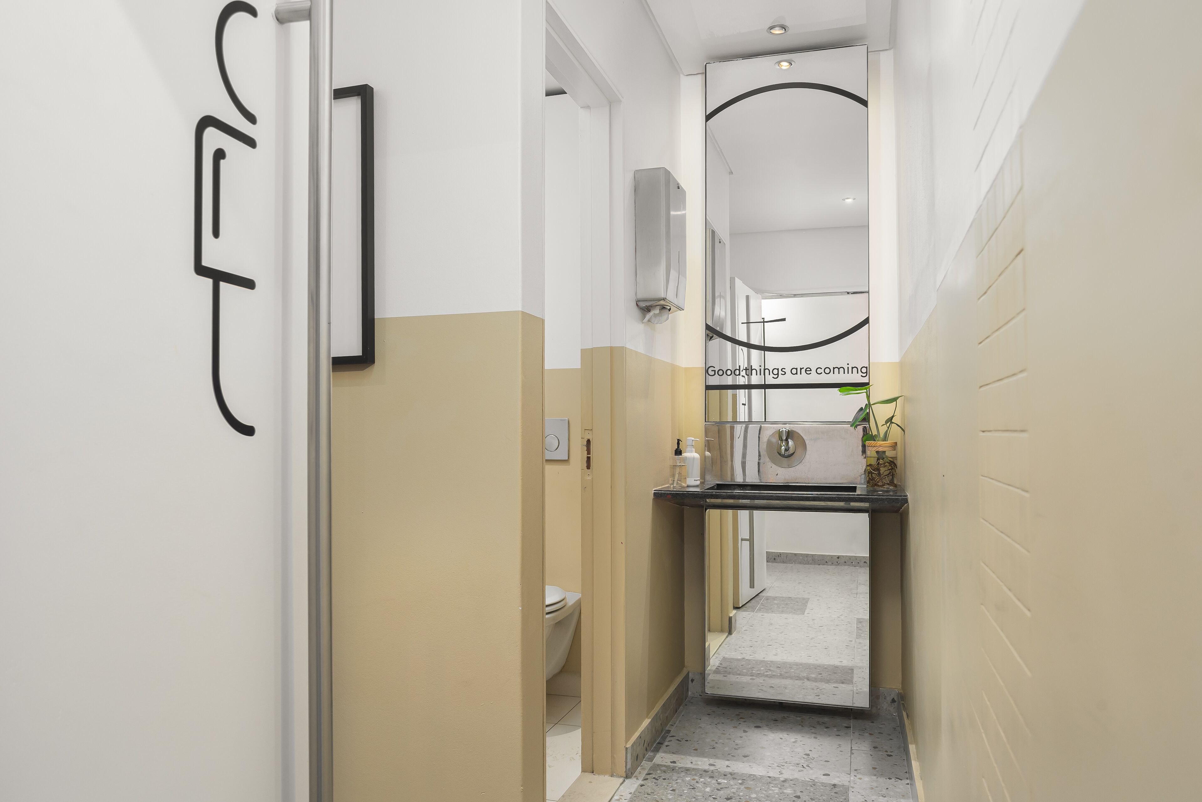 Een smalle gang met een toiletruimte en wastafel, een spiegel en een wandairconditioning.