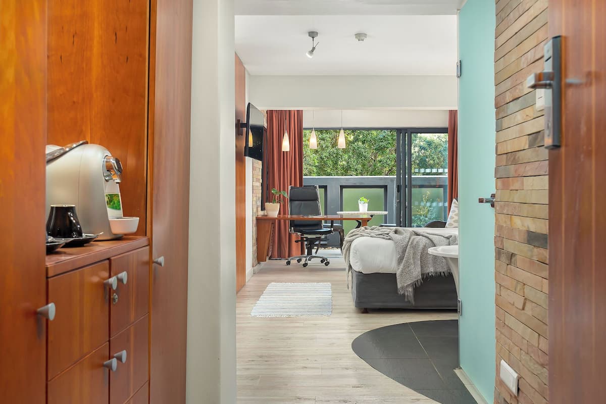 Una habitación de hotel moderna con un armario de madera, un escritorio con una silla, una cama con cabecera negra y un ventanal con vistas a zonas verdes.