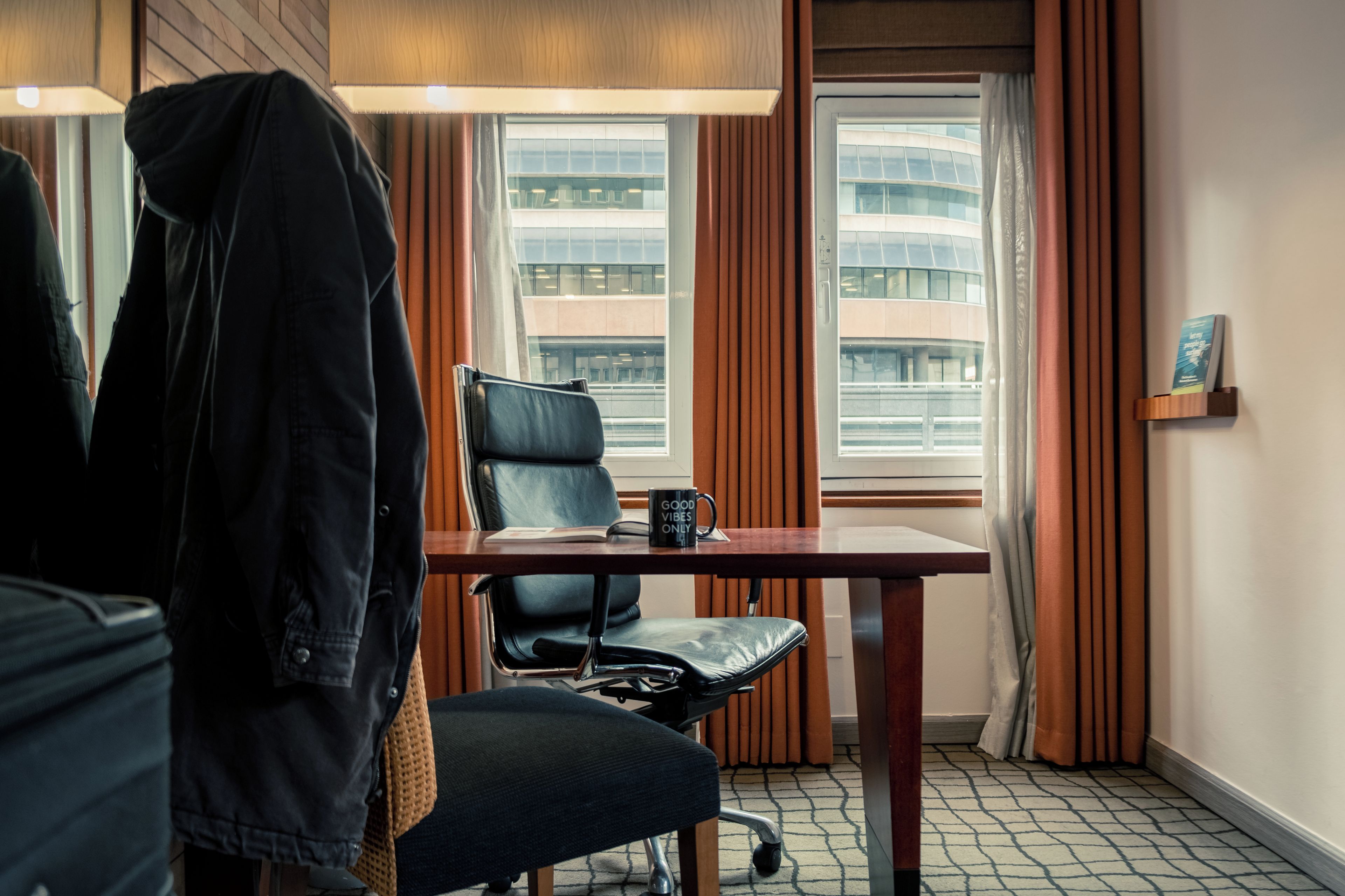 A hotel room with a desk, chair, and a jacket hanging on the wall.