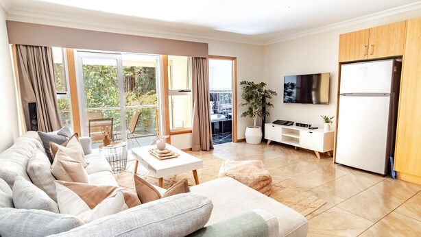 Un salon moderne avec un canapé, une table basse et un téléviseur. Il y a un balcon avec vue sur la verdure.