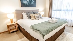 Une chambre d’hôtel bien aménagée, avec un grand lit, deux lampes de chevet, une table de chevet et une vue sur l’extérieur à travers de fines rideaux.