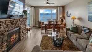 A living room with a stone fireplace, a large TV, a dining area with chairs, and a sofa with cushions. - Seasons Lakefront Escape | Pool, Hot Tub & Views (Sandpoint)
