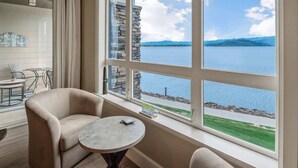 A room with a large window offering a view of a lake and mountains. There are two chairs and a small table. - Seasons Lakefront Escape | Pool, Hot Tub & Views (Sandpoint)