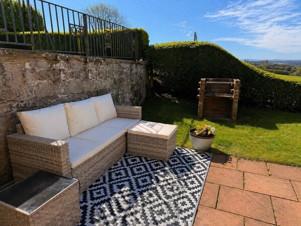 A wicker sofa with cushions, a patterned rug, a brick planter with flowers, and a stone wall.