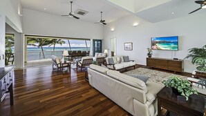 A spacious living room with a large window overlooking a beach, wooden flooring, a sofa, a dining area, and a TV mounted on the wall. - Hula House - Beachfront North Shore - NEW SPECIAL FALL RATES. (Wailua)