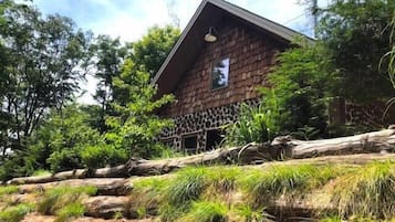 Un bâtiment rustique, doté d’un toit en pente, entouré d’une végétation luxuriante et bordé d’un terrain rocailleux.
