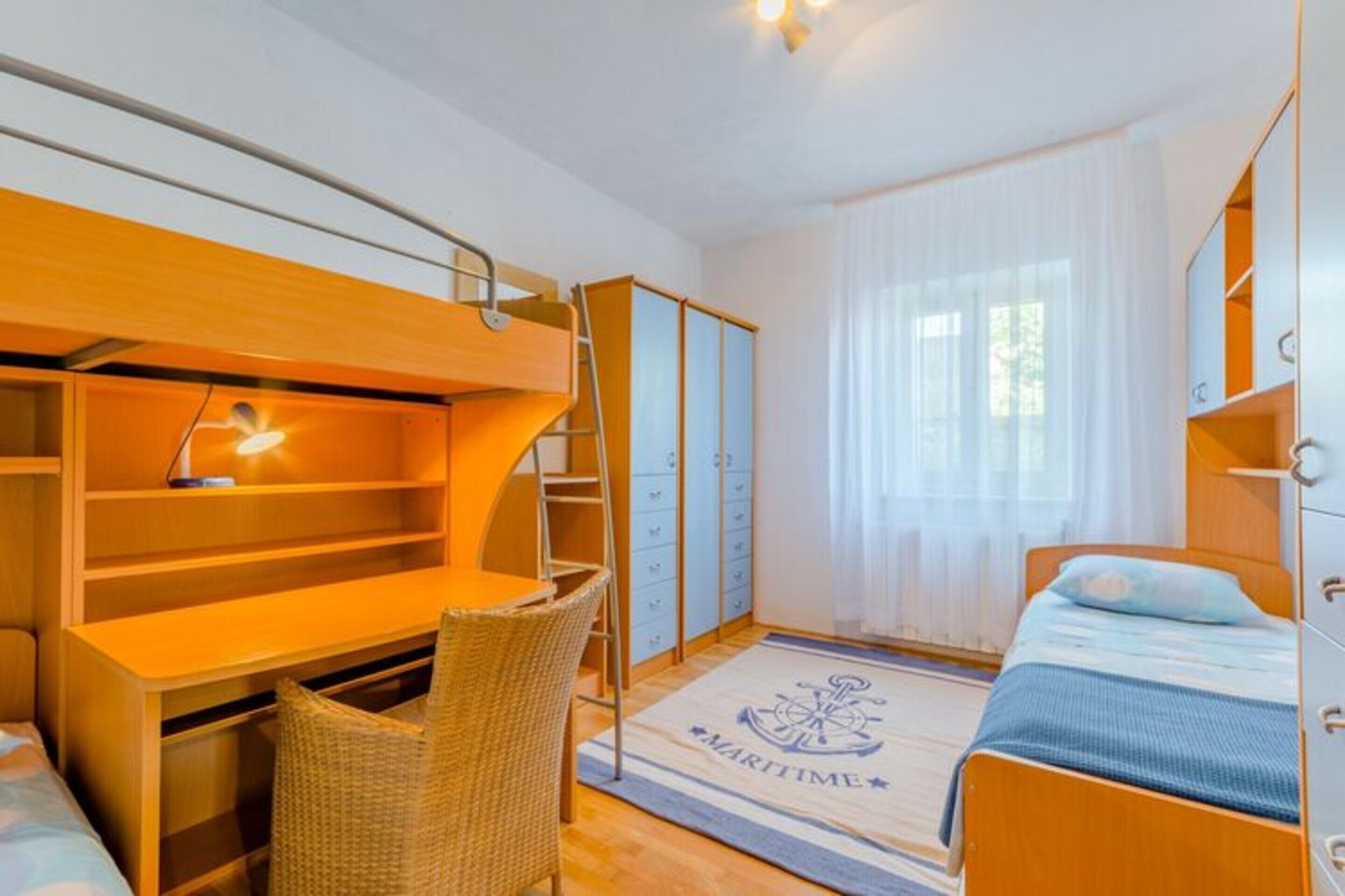 A compact bedroom with a bunk bed, a desk with a chair, a wardrobe, and a bed with a blue bedspread.
