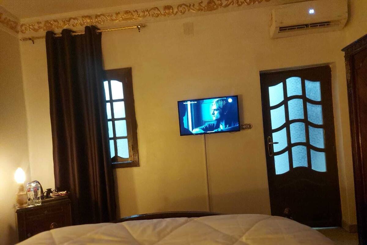 A hotel room with a bed, a television mounted on the wall, and a window with a unique design.
