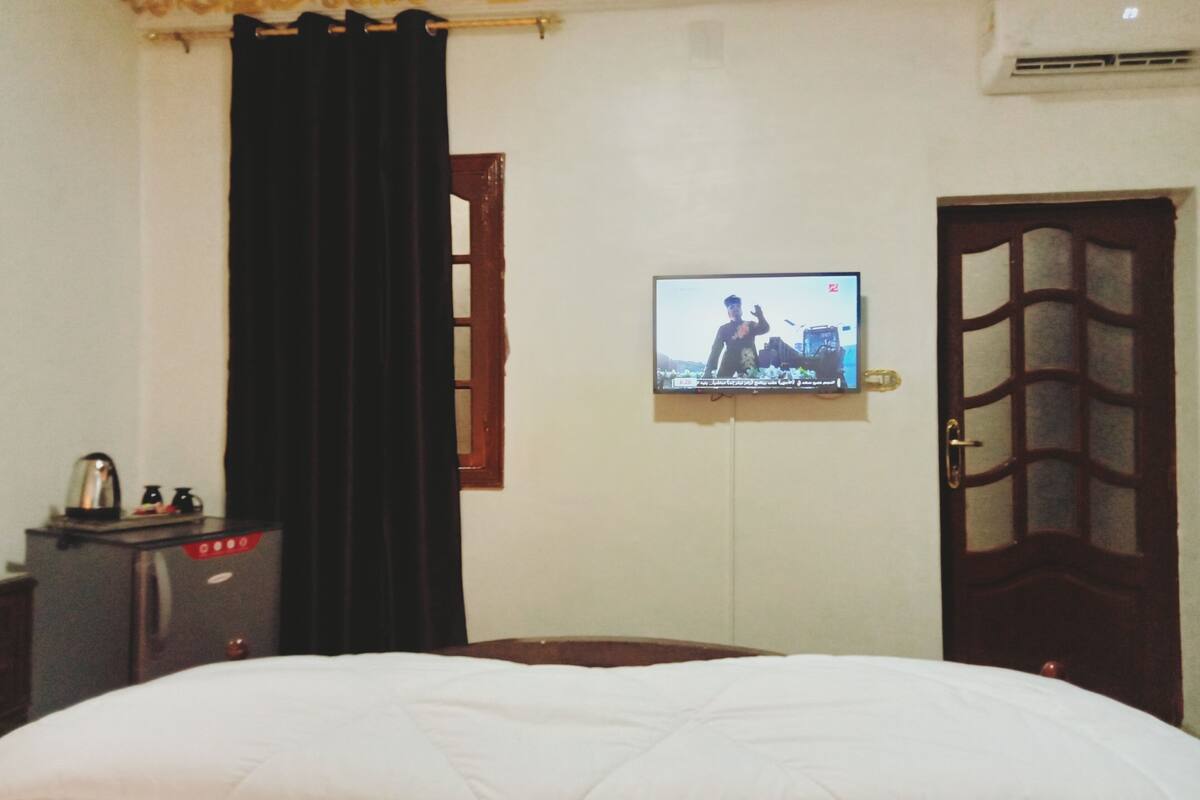 A hotel room with a bed, a television mounted on the wall, a wooden door, and a window with a curtain.