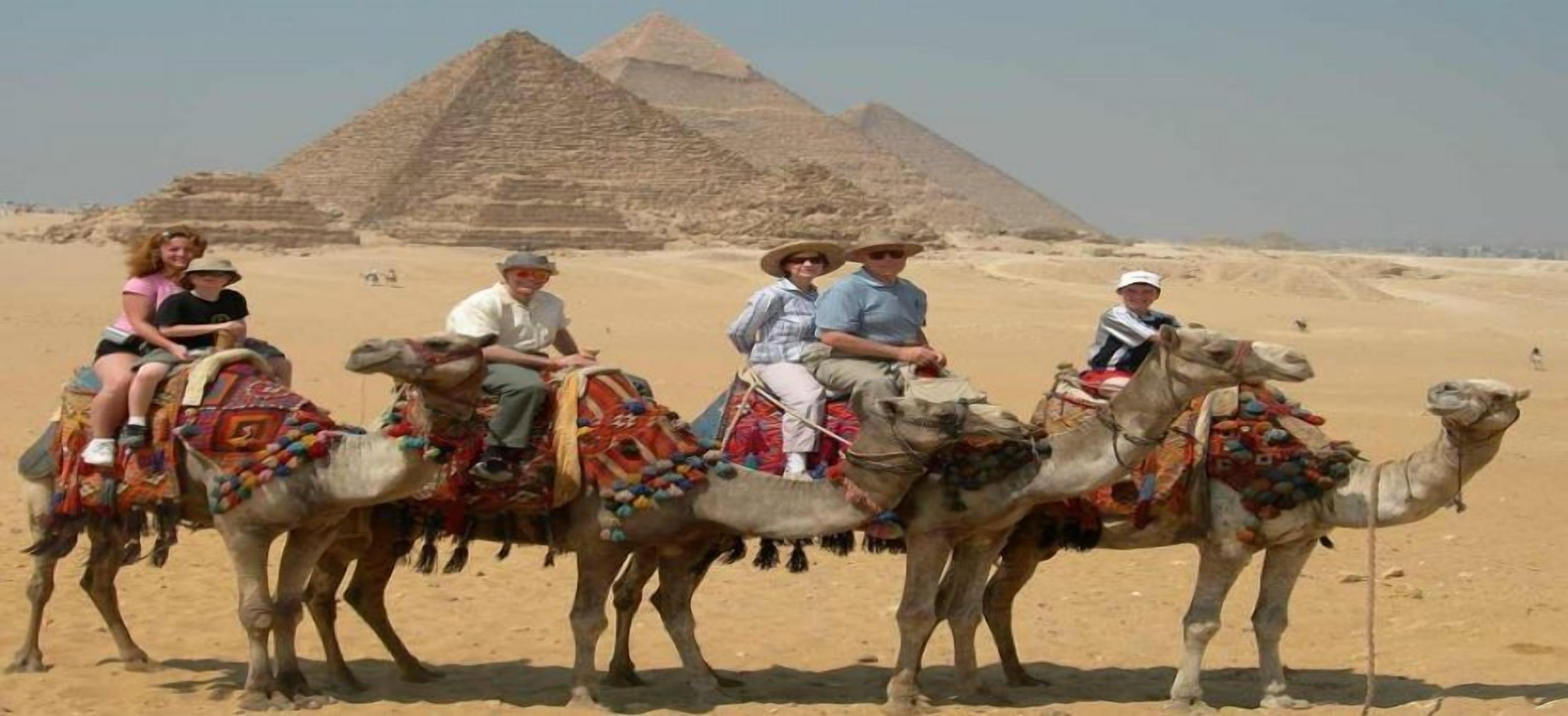 Ryhmä ihmisiä ratsastaa kameleilla aavikolla, ja taustalla näkyy pyramideja.