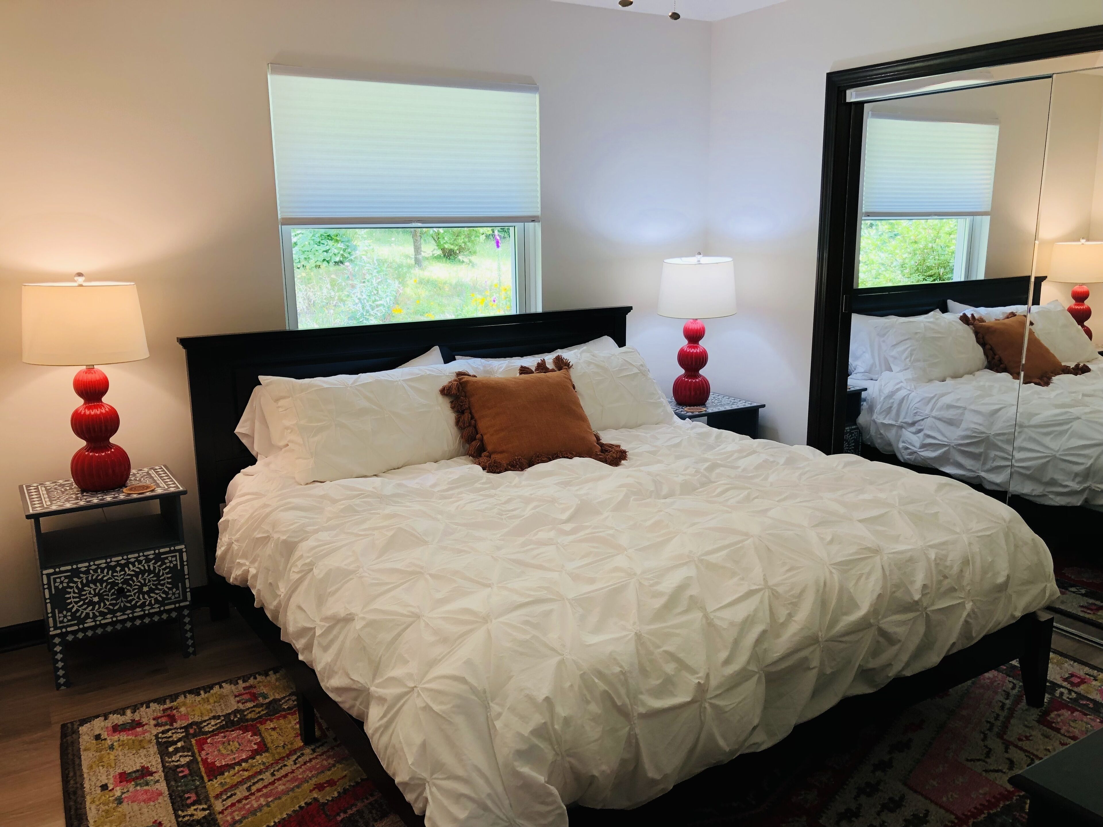 A neatly made bed with white bedding, two bedside tables with lamps, a patterned rug, and a large mirror reflecting the room.