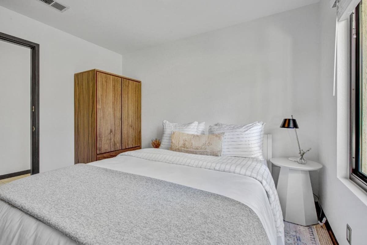 A bedroom with a bed, a wooden wardrobe, a bedside table with a lamp, and a window.