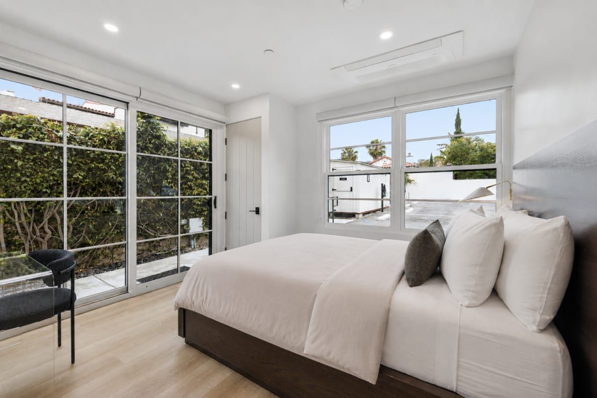 A bedroom with a large bed, a glass door leading to a balcony, and a window with a view of buildings.