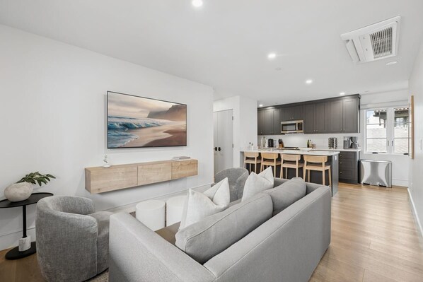 A modern living room with a grey sofa, a dining area with a table and chairs, and a kitchen with grey cabinets and a microwave.
