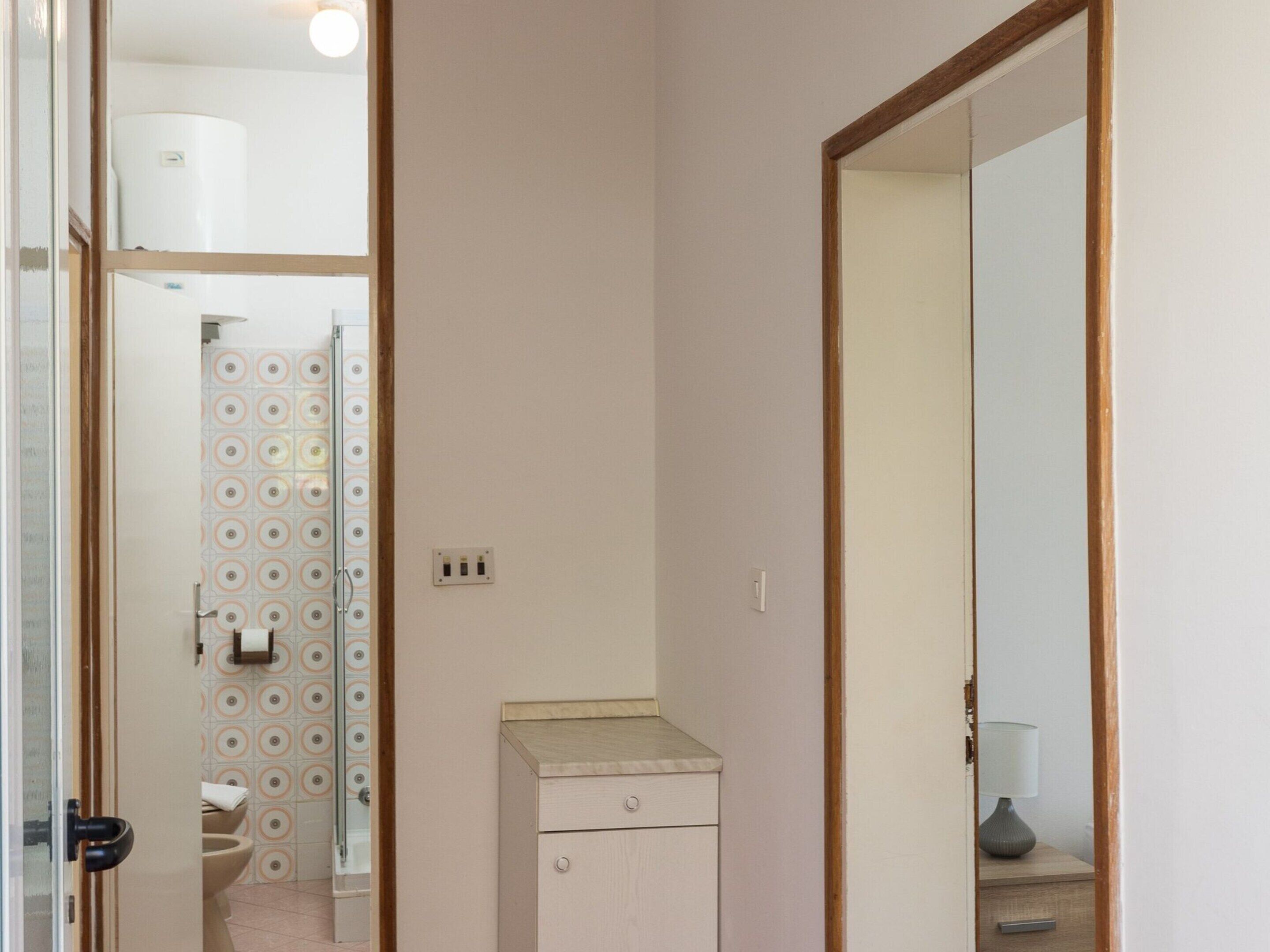 A narrow hallway with a door leading to a bathroom with a toilet and a shower. There is a small cabinet with a drawer and a mirror reflecting a lamp on a bedside table.
