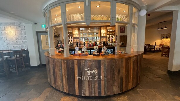 Un espace bar avec un comptoir en bois portant l’inscription « WHITE BULL », diverses bouteilles, des verres et une présentation de récompenses.