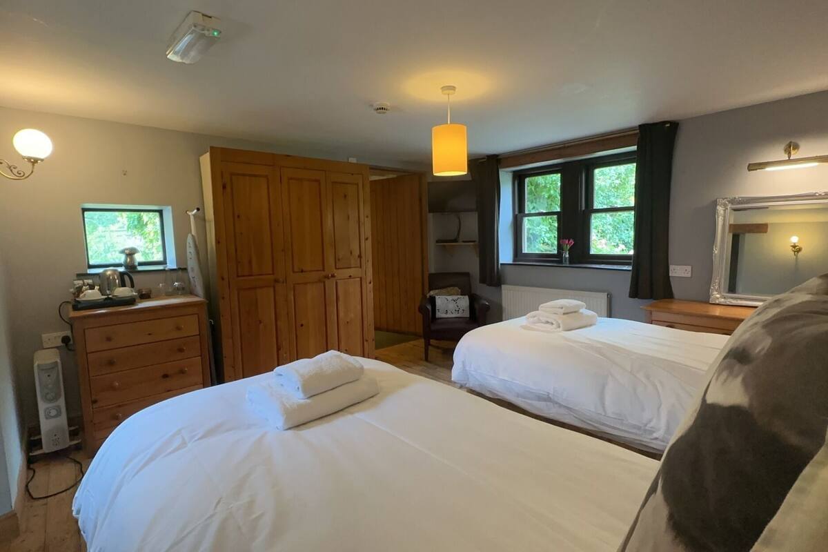 A bedroom with two beds, wooden wardrobe, bedside tables, a desk with a chair, a window with a view, and a mirror.