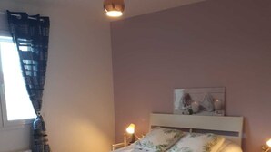 A bedroom with a bed, two bedside lamps, a window with curtains, a radiator, and a wall-mounted artwork.