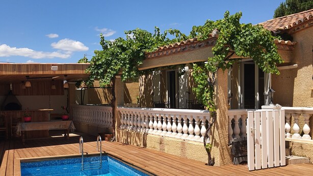 A rectangular swimming pool with a wooden deck, surrounded by a white fence and a pergola with climbing plants.