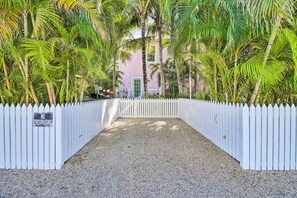 A gated entrance with a 'No Trespassing' sign, surrounded by palm trees and a white picket fence leading to a building with a pink facade.