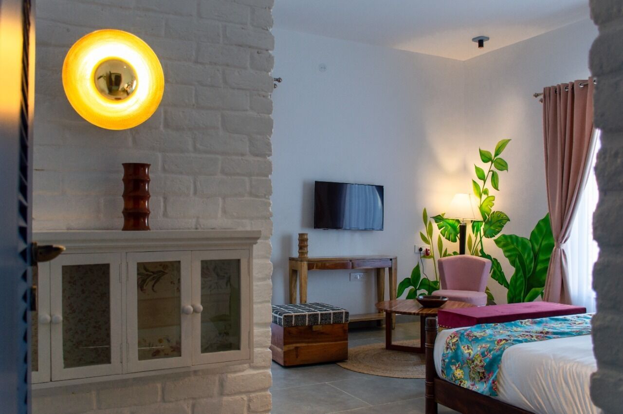 A room with a white brick wall, a television mounted above a wooden console table, a bed with a floral bedspread, a pink chair, and a large green plant.