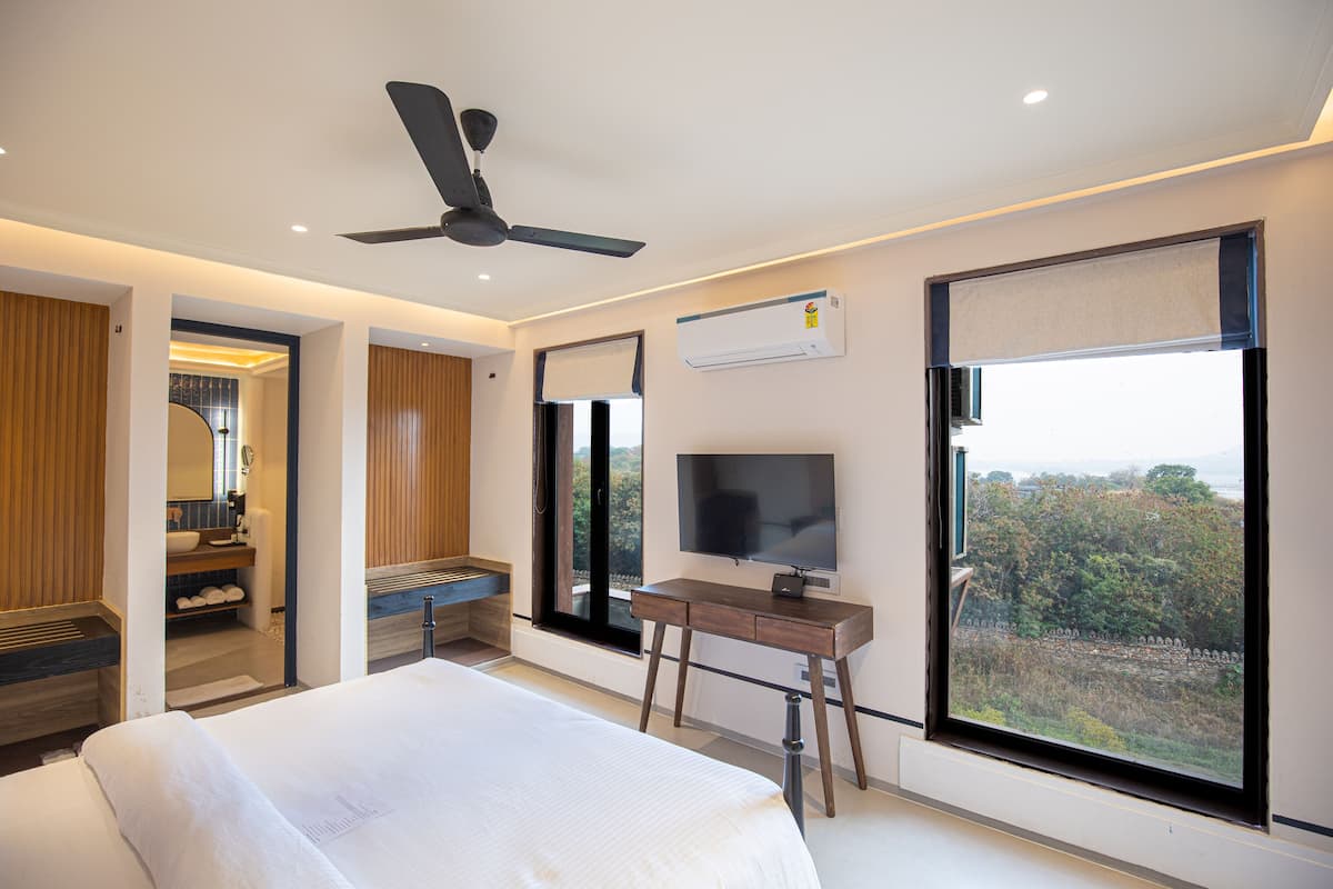 Une chambre d’hôtel moderne avec un grand lit, un téléviseur posé sur un meuble en bois, un ventilateur de plafond et une vue sur l’extérieur grâce à une grande fenêtre.