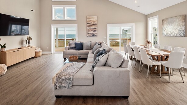 A spacious living room with a large gray sofa, a dining area with white chairs, and a view of the ocean through large windows.