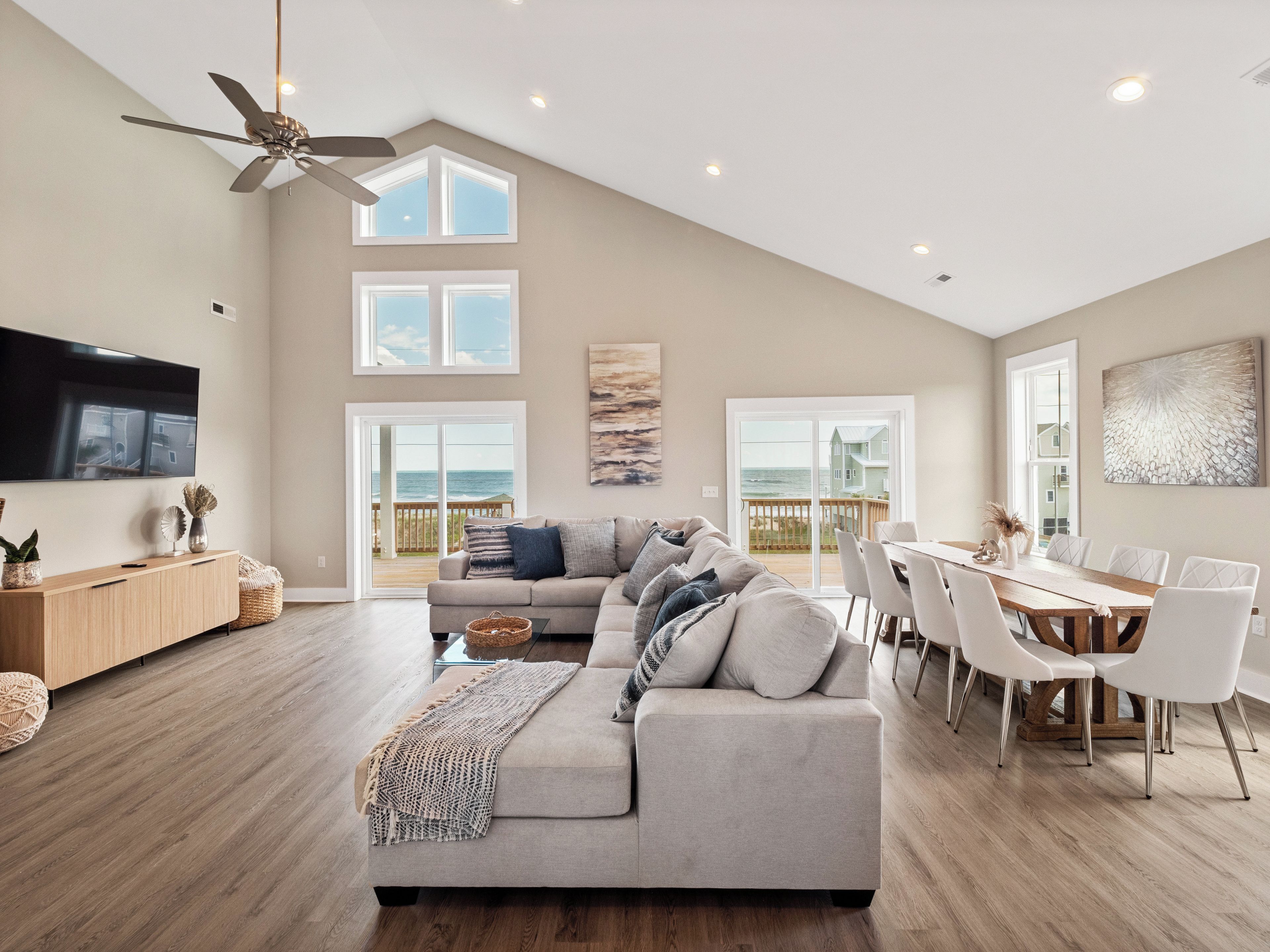 A spacious living room with a large gray sofa, a dining area with white chairs, and a view of the ocean through large windows.