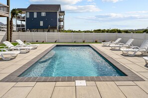 Una piscina rectangular con sillas de descanso blancas alrededor, una valla de piquete blanca y un edificio azul al fondo.