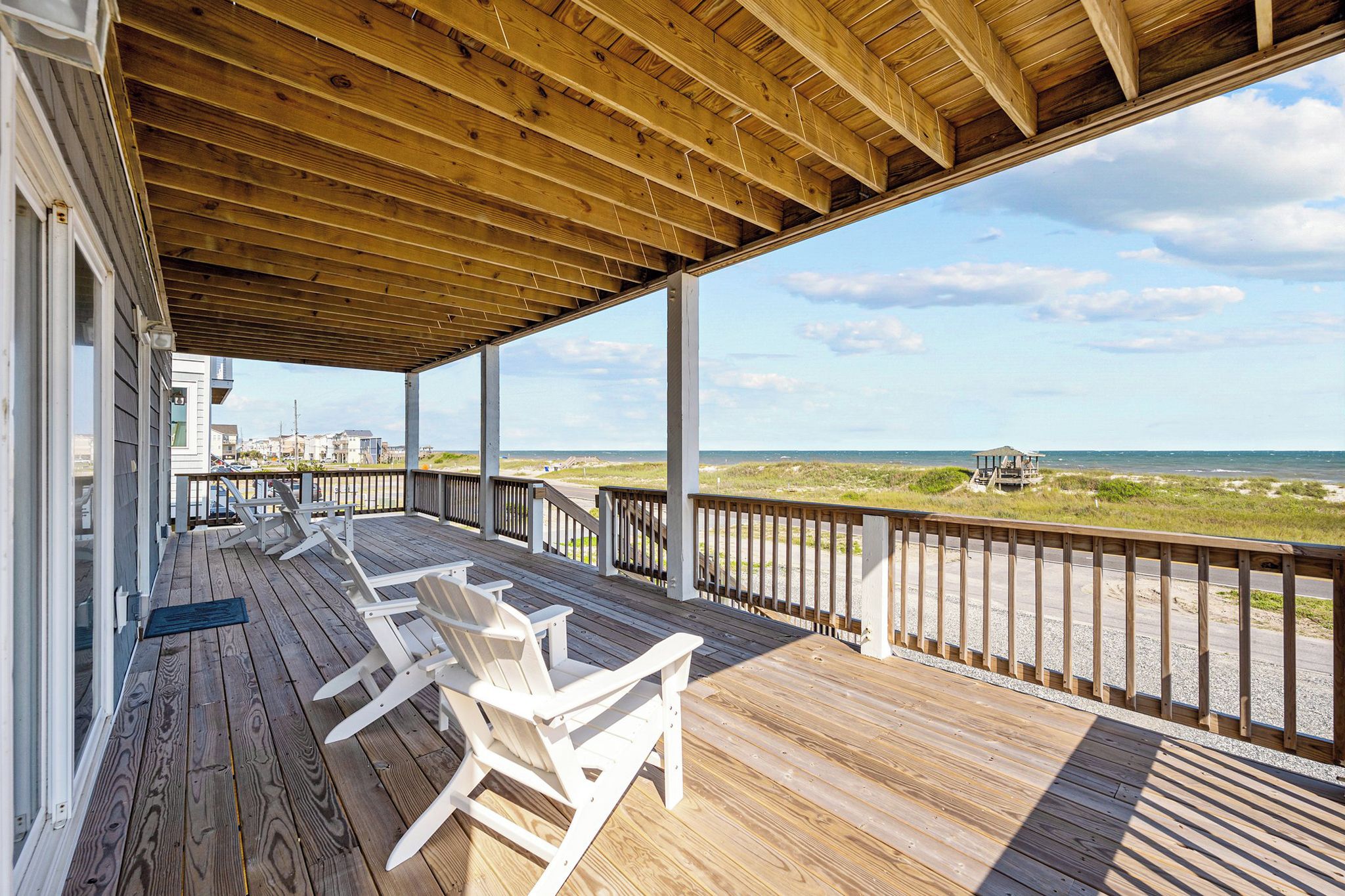 A wooden deck with white chairs, a wooden railing, and a view of the beach and ocean.